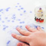 A hand is placed on top of the slime that has snowflake confetti.The snow globe is to the right behind the slime with more snowflake confetti to its left. The text is placed at the top of the image saying "winter slime recipe"