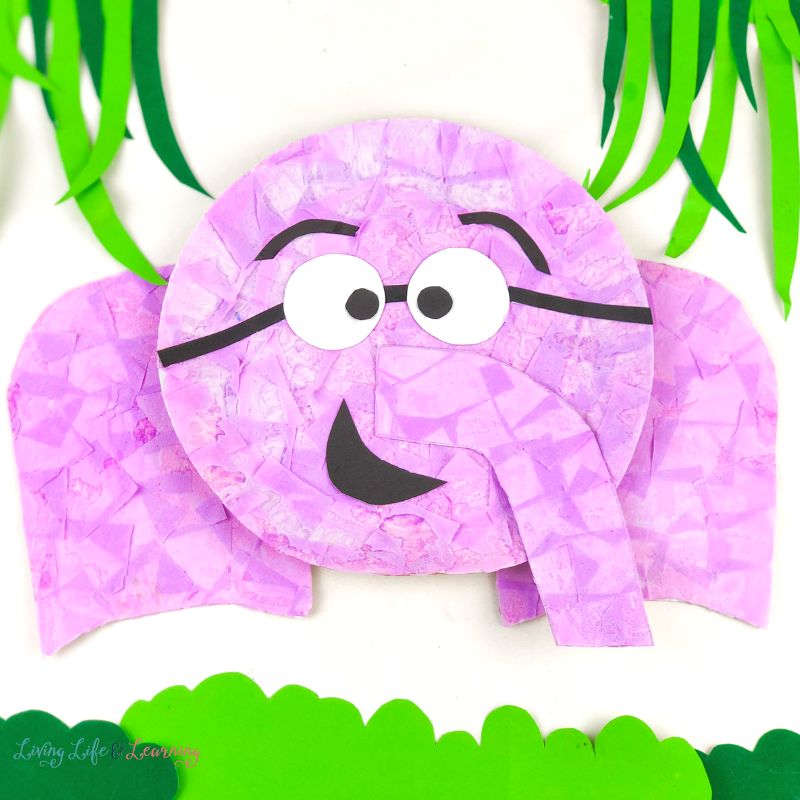 Pink paper plate elephant with green DIY grass and bush surrounding it on a white background.