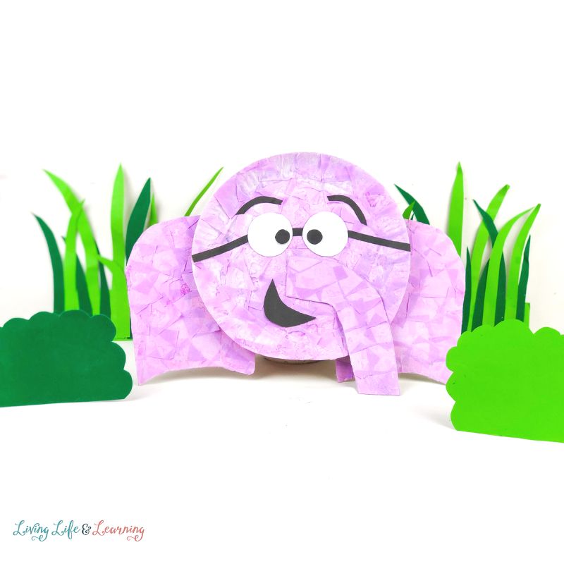 Pink paper plate elephant in front of green DIY grass and bushes.
