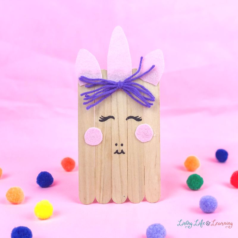 Wooden unicorn craft made of popsicles in a pink background surrounded by colorful fuzzy balls.
