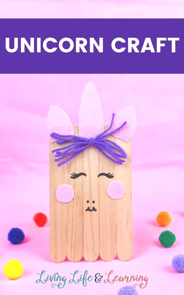 Wooden unicorn craft made of popsicles in a pink background surrounded by colorful fuzzy balls.